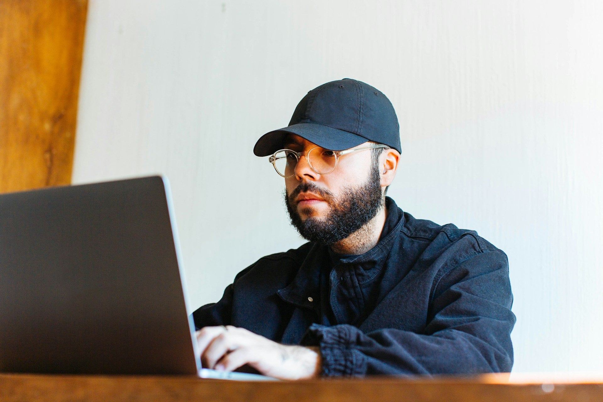 Man wearing glasses and a dark cap concentrating on a laptop